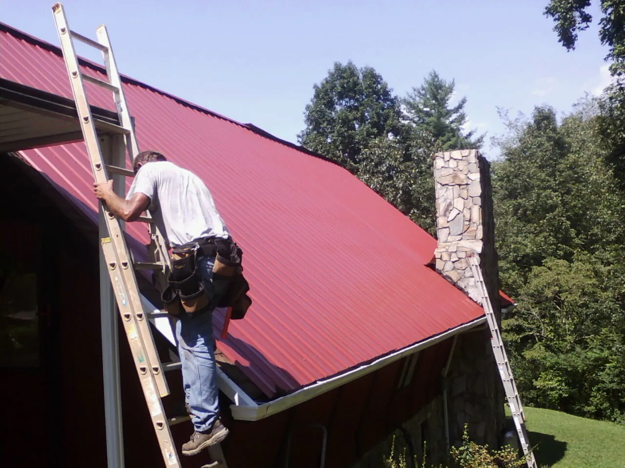 Roofer on a ladder beside a red metal roof and stone chimney — Soffit & Fascia Repair by Roofing Brothers in Owings